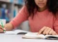 Estudante revisa conteúdo com livro, caderno e notebook enquanto faz uma pausa para o lanche.