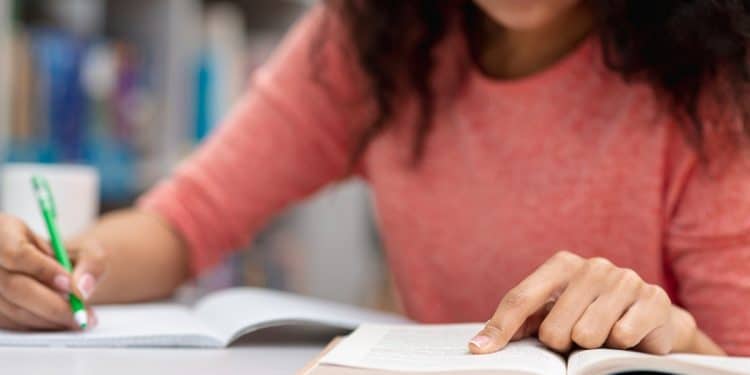 Estudante revisa conteúdo com livro, caderno e notebook enquanto faz uma pausa para o lanche.