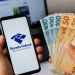 Celular com acesso à Receita Federal ao lado de notas de dinheiro representando dados de renda média dos trabalhadores brasileiros.