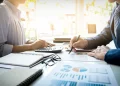 Profissionais analisam gráficos e dados financeiros em uma mesa de trabalho com calculadora e documentos.