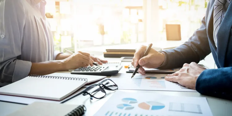 Profissionais analisam gráficos e dados financeiros em uma mesa de trabalho com calculadora e documentos.