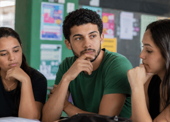 Três estudantes com caderno sobre a mesa em área comum de faculdade, em momento de escolha do curso.
