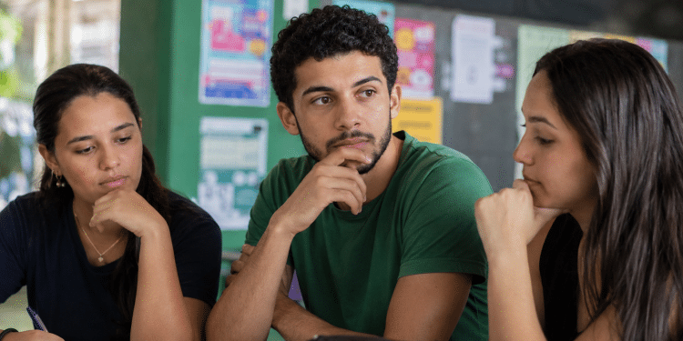 Três estudantes com caderno sobre a mesa em área comum de faculdade, em momento de escolha do curso.