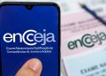 Celular exibindo o logotipo do Encceja, com folhas de prova desfocadas ao fundo