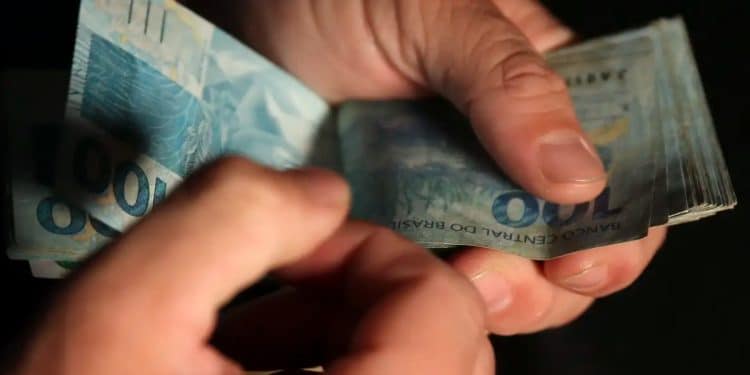Mãos contam notas de R$ 100, referência ao piso de R$ 3.242 pago em 2026 ao técnico em radiologia, valor equivalente a dois salários mínimos.