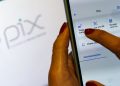 Mão feminina com unhas vermelhas usa aplicativo de banco em smartphone na tela Pix, logo do Pix ao fundo, simbolizando a restituição automática do IR de até R$ 1.000.