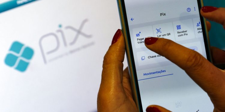 Mão feminina com unhas vermelhas usa aplicativo de banco em smartphone na tela Pix, logo do Pix ao fundo, simbolizando a restituição automática do IR de até R$ 1.000.
