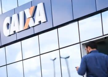 Homem mexe no celular em frente à fachada de uma agência da CAIXA, banco que libera o Saque Calamidade do FGTS para novas cidades