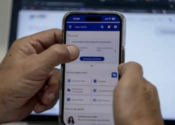 Pessoa segurando celular com a tela inicial do aplicativo Meu INSS aberta, exibindo os atalhos de Prova de Vida, Extratos, Empréstimo Consignado, Consultar Pedidos e Novo Pedido, que dão acesso aos serviços resolvidos sem ida à agência