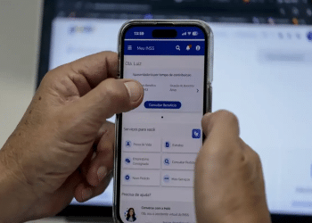 Mãos segurando celular aberto no app Meu INSS com tela de serviços e botão “Consultar Benefício”, simbolizando os serviços do Meu INSS.
