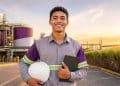 Jovem aprendiz sorridente, de uniforme cinza e roxo com faixas refletivas, segura capacete branco e tablet em frente a tanques e canavial de usina da Raízen, empresa que recebe inscrições para o Programa Aprendiz 2026 até 27 de julho